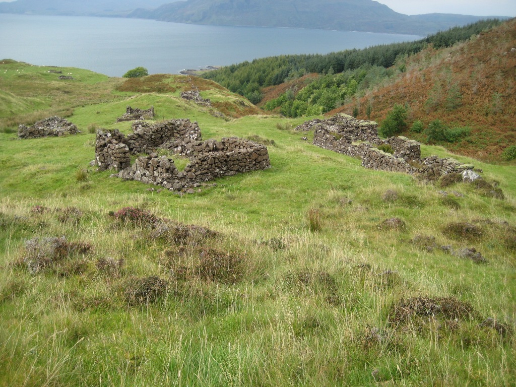 ruined remains of several buildings on a grassy slope overlooking a body of water 