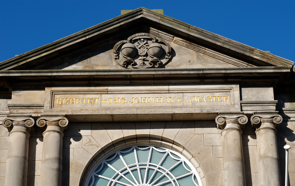 A close-up photo of the facade of Trinity House in Leith. It shows the top of the buidling which features an ornate semi-circular window along with stone carvings.