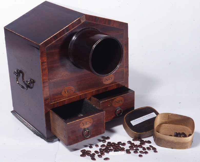 A small mahogany box with a central opening above two small drawers which are marked "yes" and "no". There are seeds pictured beside the box.
