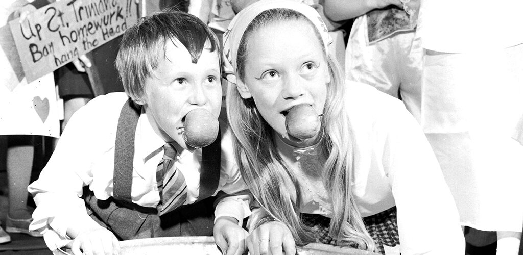 A young boy and girl dooking for apples. The crouch over a basin full of water, their faces and hair wet, each with an apple between their teeth.