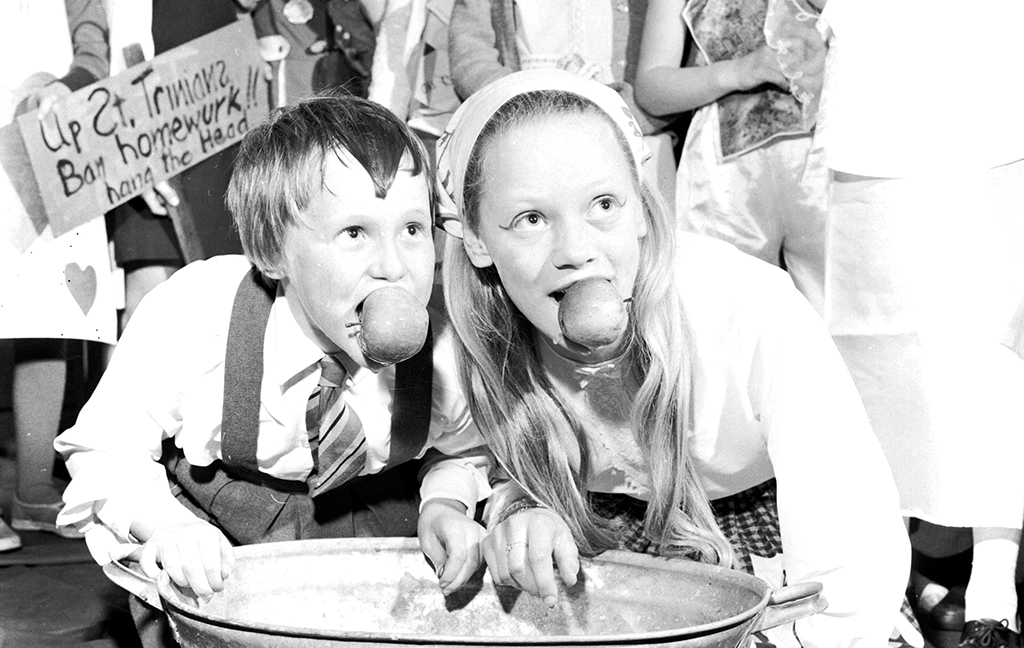 A young boy and girl dooking for apples. The crouch over a basin full of water, their faces and hair wet, each with an apple between their teeth.