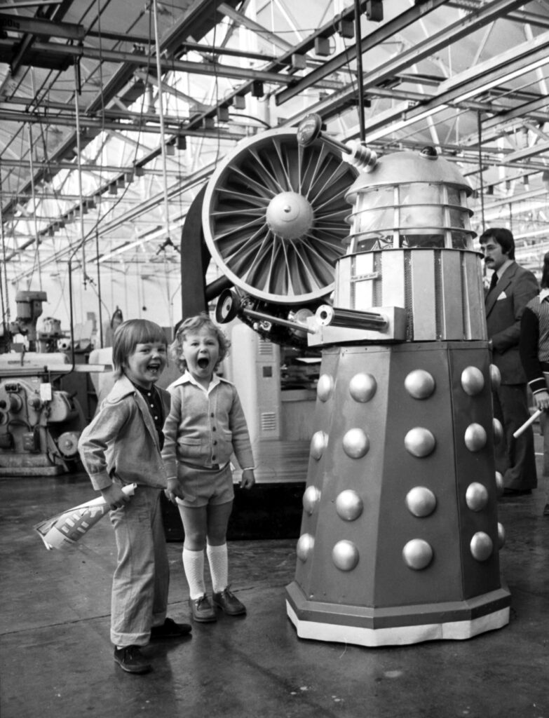 A black and white archive photo showing teo young children looking at a Dalek smiling.