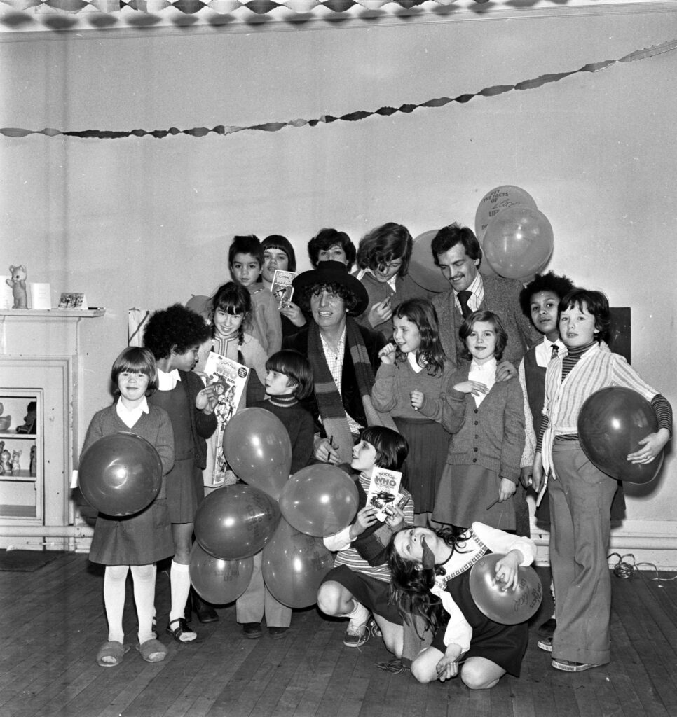 A black and white archive photo of Tom Baker surrounded by children holding balloons.