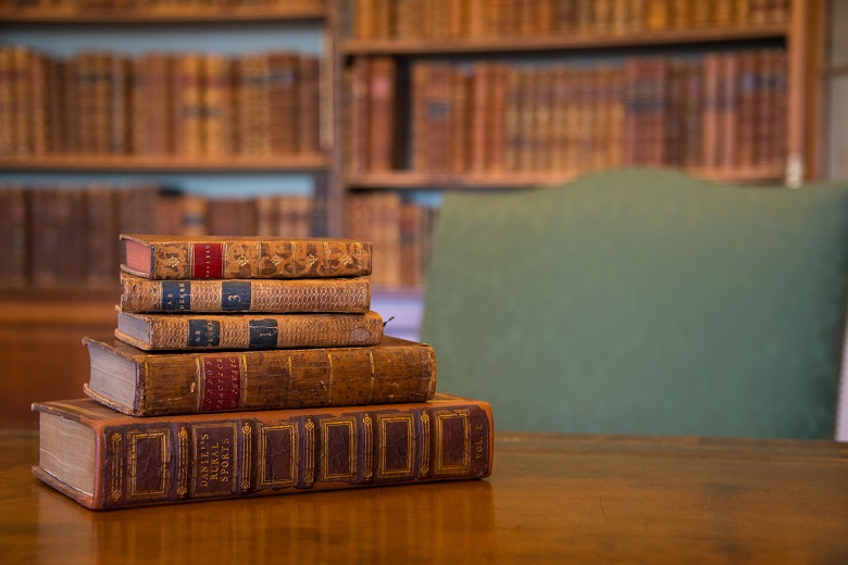 A pile of five old leather-bound books sitting on a table. In the background, there are library shelves filled with antique books.