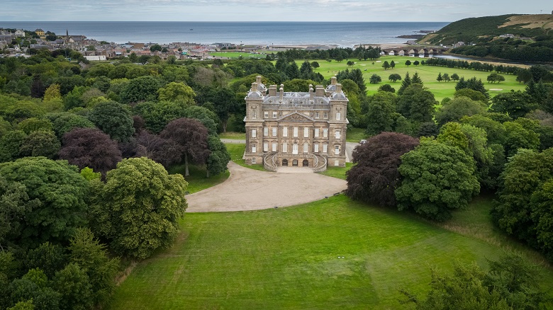 Aerial image of Duff House, a large Georgian mansion set in parkland with a large gravel driveway in front. In the distant background is the sea and a seaside village.