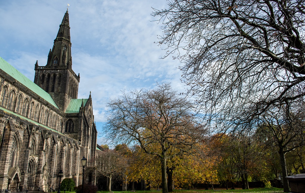 Autumnal trees in front of Glasgow Cathedral, a large and imposing stone building with a grene roof and a tall spire.