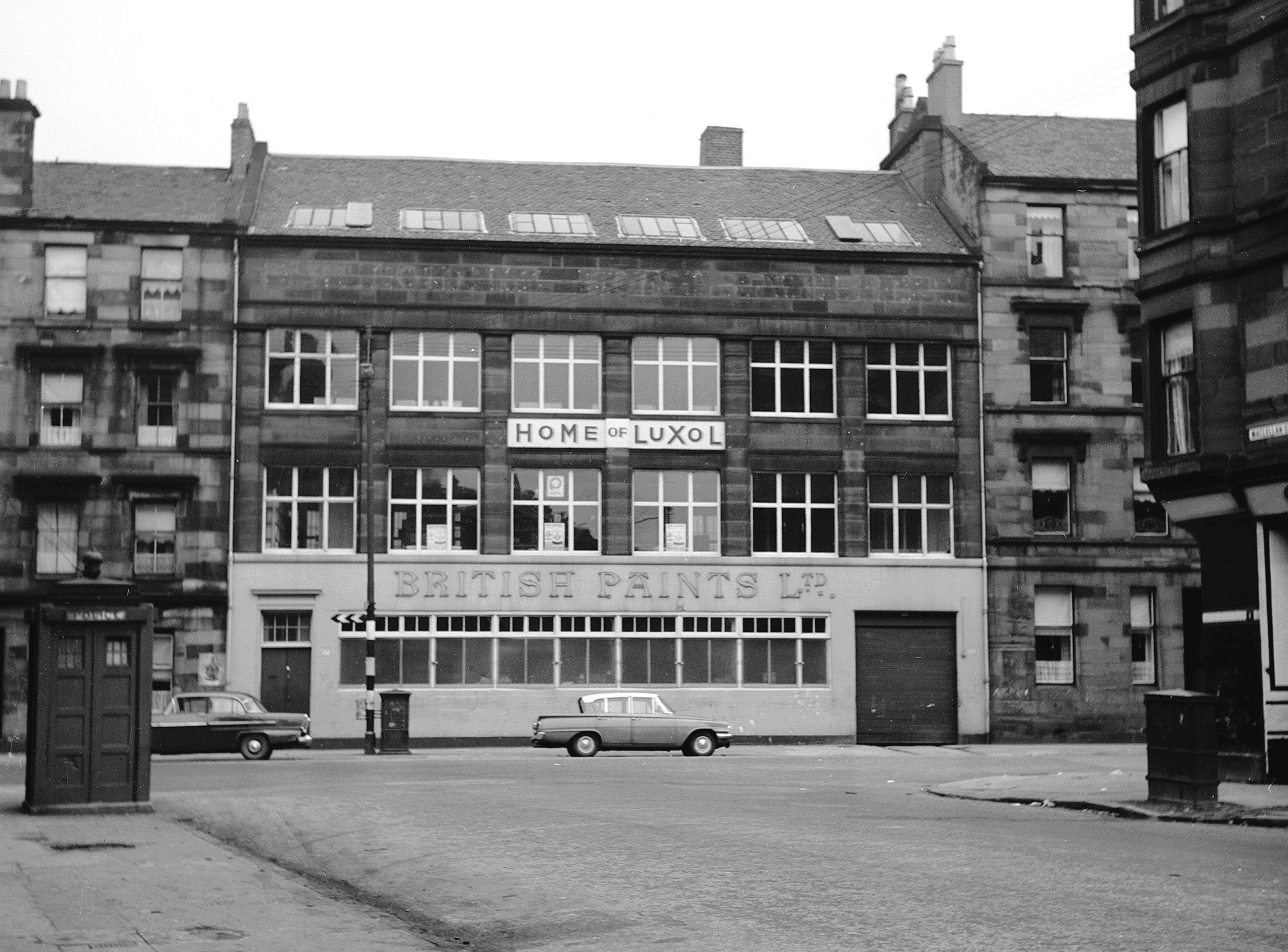 A grainy black and white archive photo from Glasgow in the 60s showing a building with a big sign "British Paints Ltf", some cars and a police box.