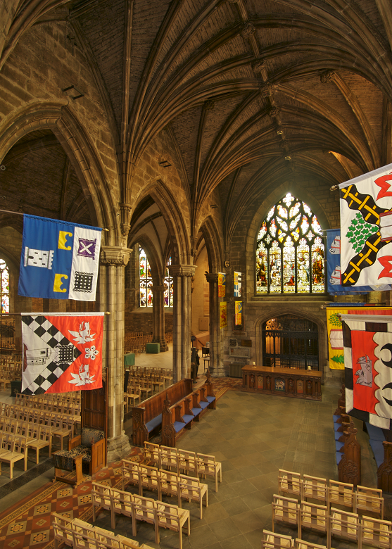 The interior of a historic cathedral with many rows of chairs centred on a dark wood altar. Light streams in through a large stained glass window and various histroic flags and standards hang from the walls. 