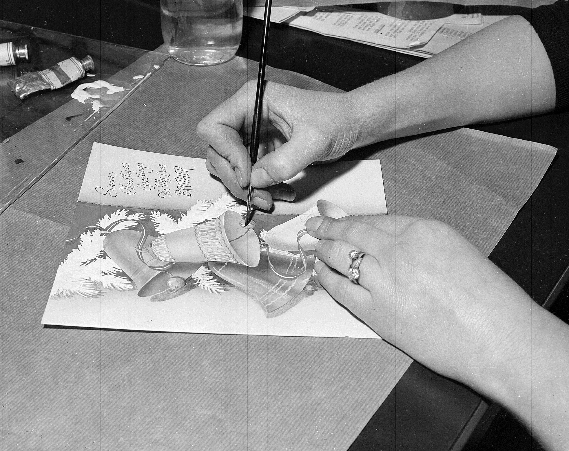 Black and white archive photo. A close up of hands illustrating an almost finished Christmas card with bells on it. 