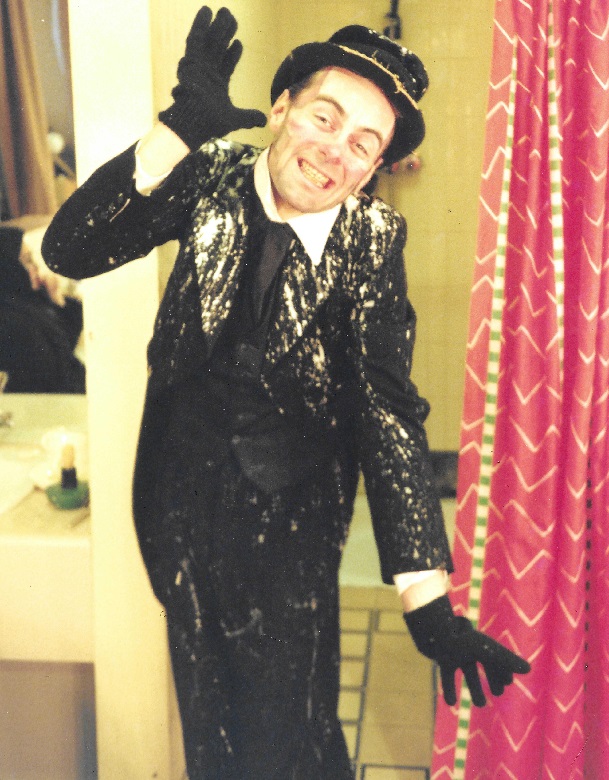 A scanned photo of an actor posing backstage in pantomime costume, The costume consists of a black suit, black hat and black gloves, all of which have been covered in a white liquid, perhaps paint. 