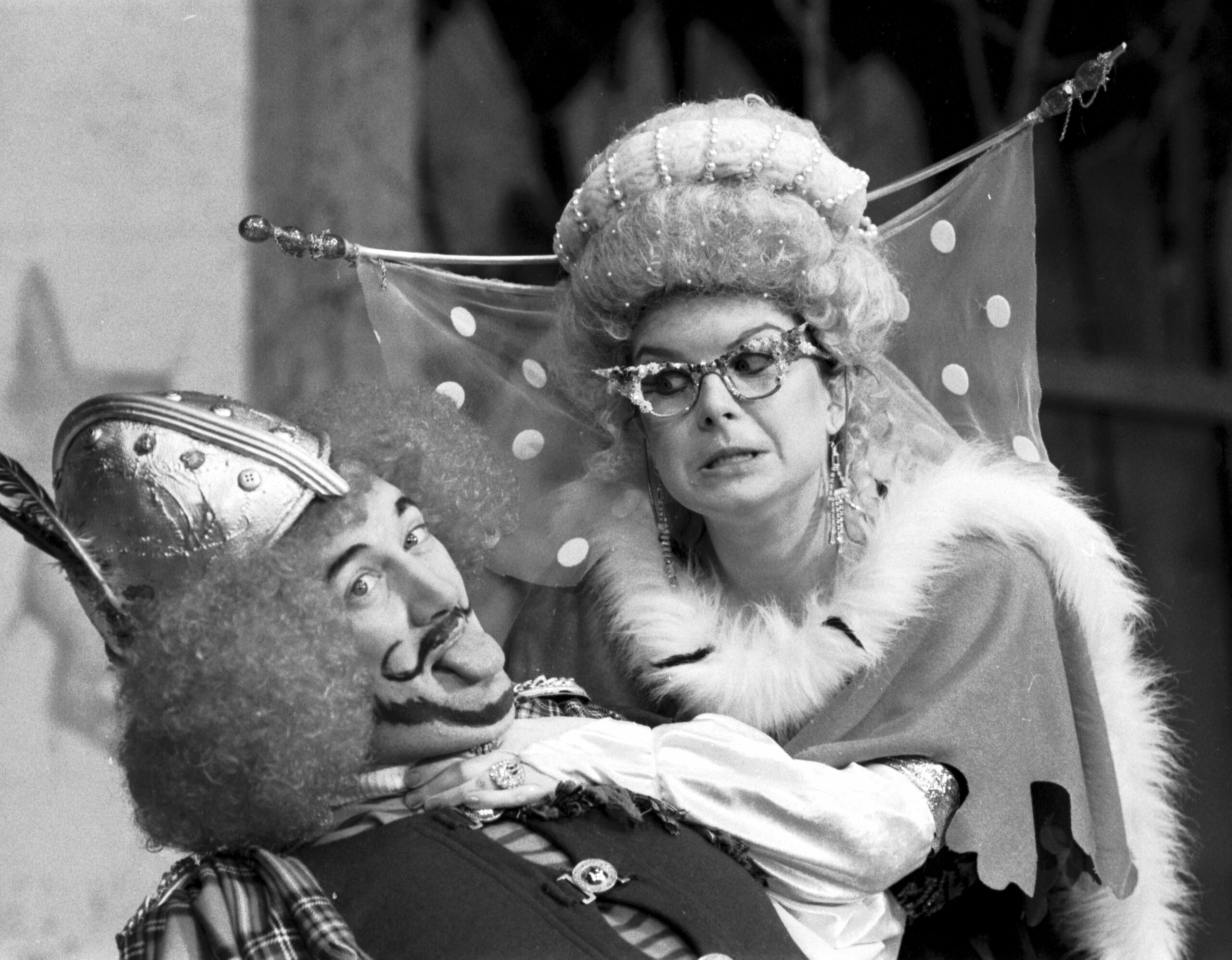 A black and white photo of Elain Smith and a co-star mid-scene during a pantomime. One appears to have some sort of medieval costume featuring a metal helmet, a large wig and a comedy moustache. Elaine C Smith is wearing a fur-lined dress, large glasses and extravagant headwear.