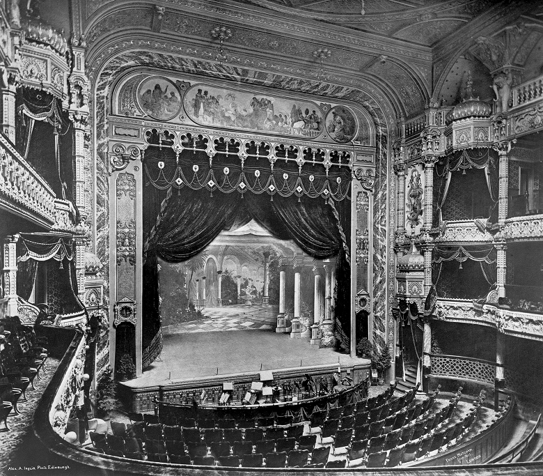 A vintage photograpah of the lavish interior of a Victorian theatre. The stalls are flanked by royal boxes and there is an orchestra pit in front of the stage. Classical paintings adorn the walls around the stage and chandeliers hang from the high ceiling. 