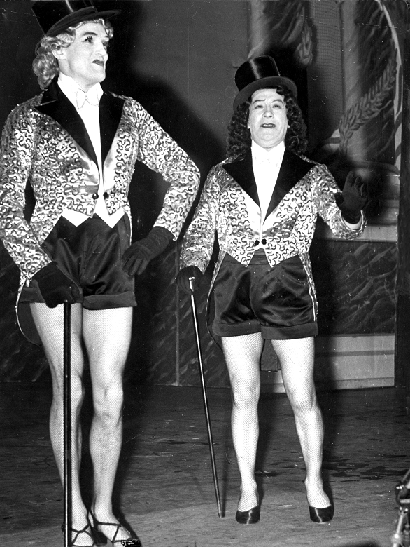 A black and white archive photo of two male actors on stage in top hats, high heels and sparkly gold jackets. Each hold a black cane and appear to be midway through a comedy or dance routine. 
