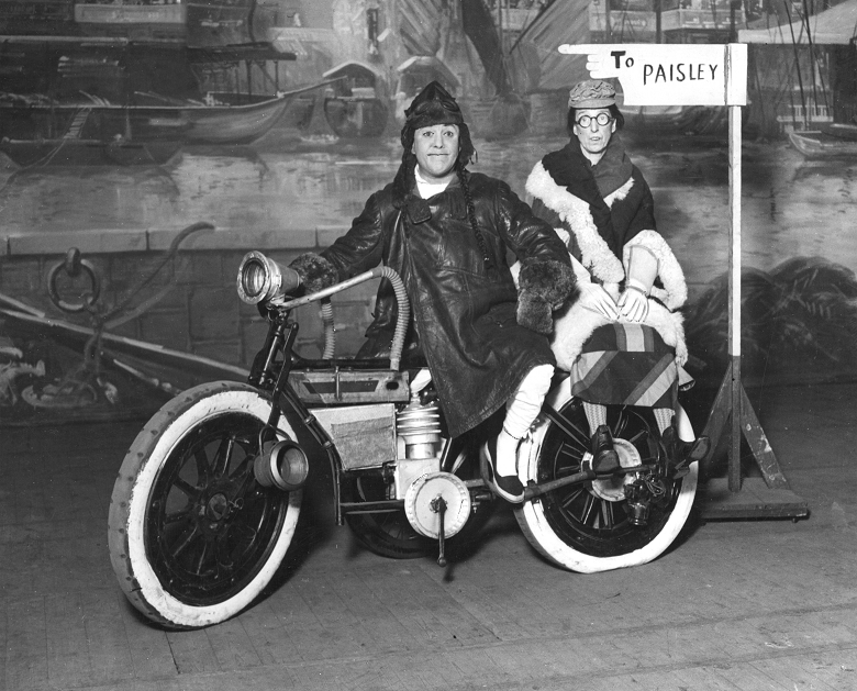 Two pantomime performers on stage with a prop motorbike. The scenery behind them suggests the scene is taking place at a docks or beside a river. A wooden fingerpost is pointing the direction to Paisley.