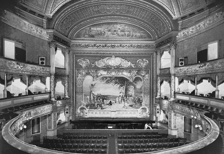 An archive photo of the interior of a theatre. The stalls, circle and royal boxes from a horseshoe shape around the stage which is framed by lavish, classical paintings and decorations. 