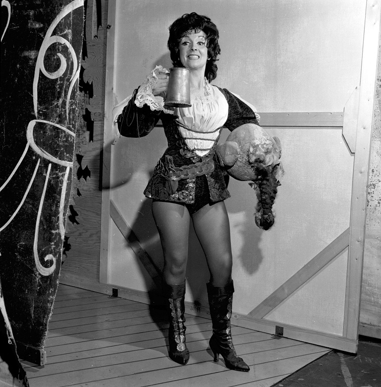 An actress poses in a typical costume for a pantomime's pricnipal boy: a shirt, jacket, tights and thigh boots. She is holding a large metal tankard and a feathered hat. 