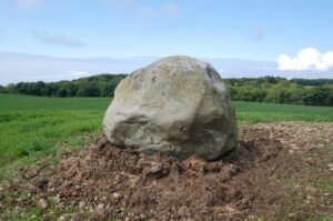 A large round boulder in a field. The boulder is 2m tall by 7m across and is light grey in colour.