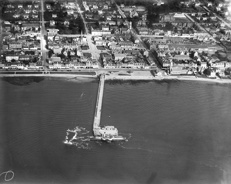 A black and white aerial photo of the topwn of Helensburgh. Shops and houses, along with a church, line the seafront. In the middle of the image there is a lengthy and grand pier. Resiential housing and train lines are in the background. 