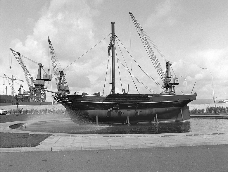 An archive black and white photo of a replica of the Comet steamboat. It stands in a small, shallow artificial lake at the edge of a paved road. In the background are the cranes and buildings of a shipyard. 