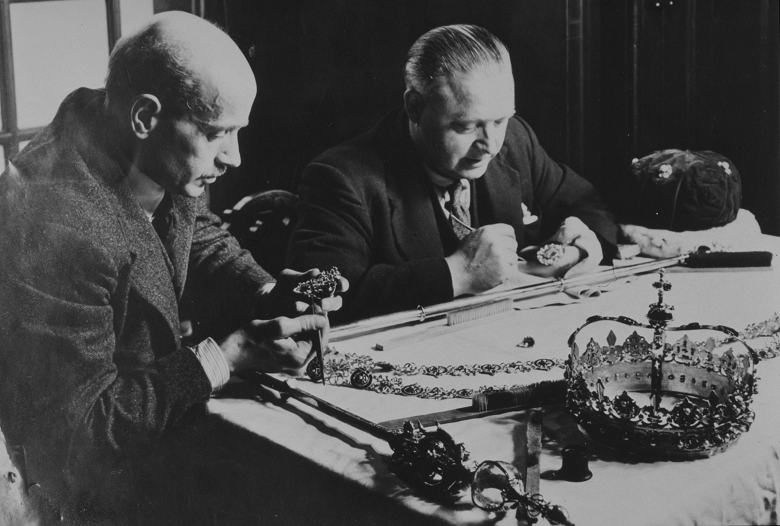 Two men in typical 1950s woollen suits sit at a table handling the crown, sceptre and other regalia. Both hold small paint brushes for carrying out intricate work.