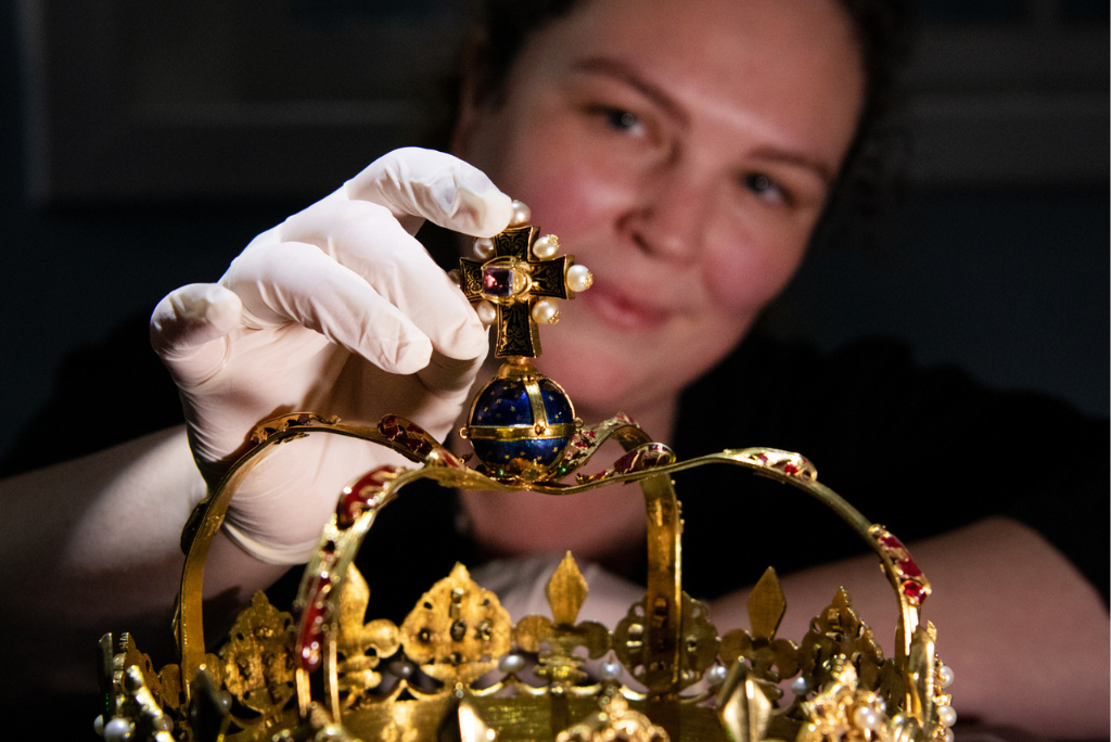 A close up of the Crown of Scotland. A woman's face is out of focus behind it. She reaches a gloved hand to adjust a decorative orb on top of the crown.