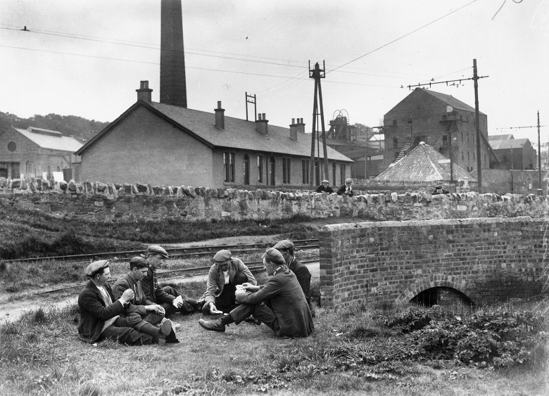 A group of six men sit in a circle on the ground outside a colliery. They are wearing working class clothing of the period (1926), including flat caps and tweed jackets.