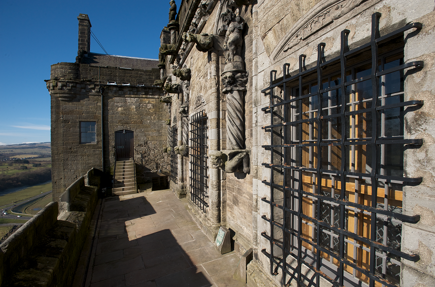 An open walkway leads to some steps and a door. On the left is a view over the landscape. On the right is a castle wall with barred windows and carved decoration.