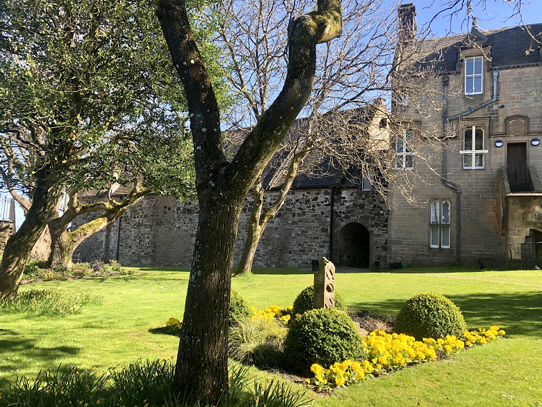 A peaceful garden within the grounds of Stirling Castle. A neat lawn interspaced with snowdrops, flower beds and old trees is flanked by stone castle buildings. 