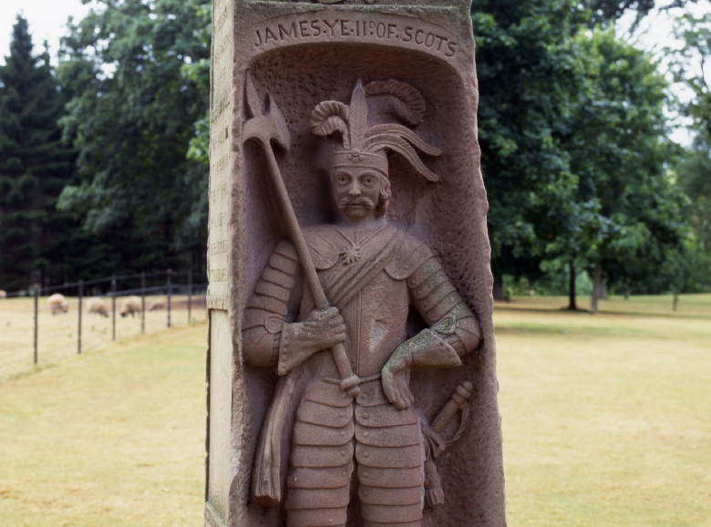A close-up photo of an obelisk featuring a carved stone statue of James II. He is depicted wearing armour and an extravagant hat decorated with feathers.