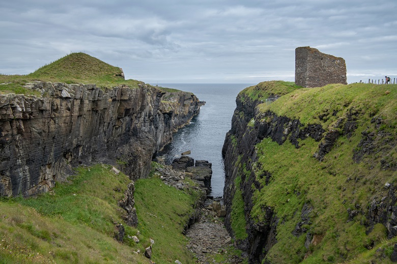 A narrow coastal inlet with steep cliffs on each side. On one side of the gap there is a ruined castle tower.