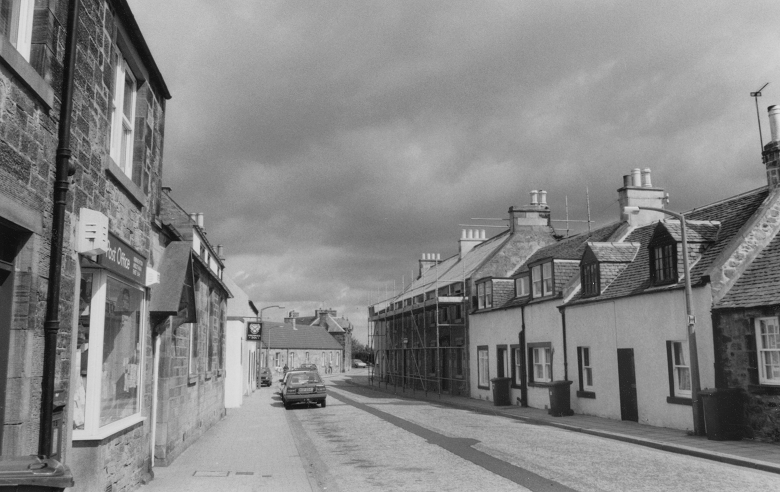 A typical terraced high street in West Lothian with small two storey cottages with dormor windows. The post office and the local pub are on the left hand side of the street and one of the buildings has scaffolding.