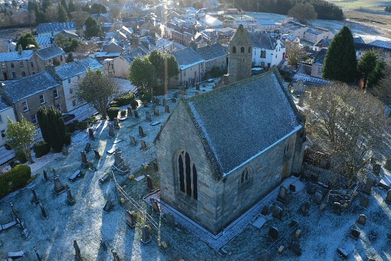 An aerial view of a modest stone church on a snowy day. It is surrounded by tombstones and nestles among more modern village houses. 