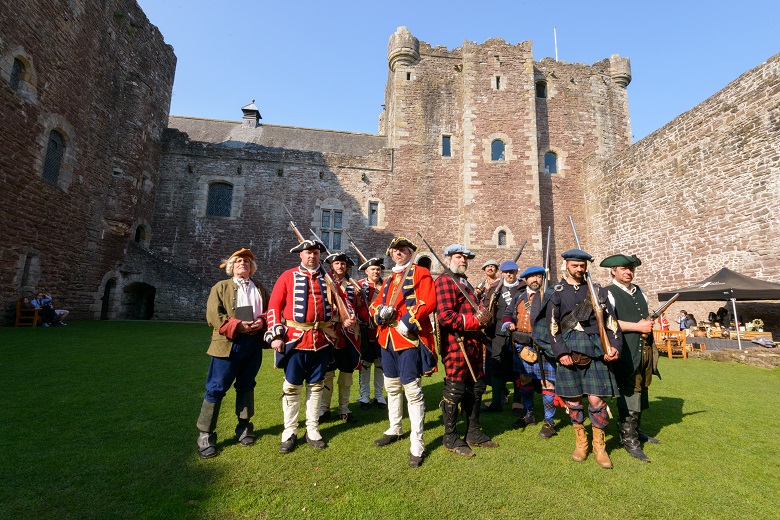 A group of historical reenactors pose in costume in front of a castle. Some are dressed as Redcoat soldiers with muskets, others are Jacobites in kilts. One man in a brown jacket holding a book represents John Witherspoon.