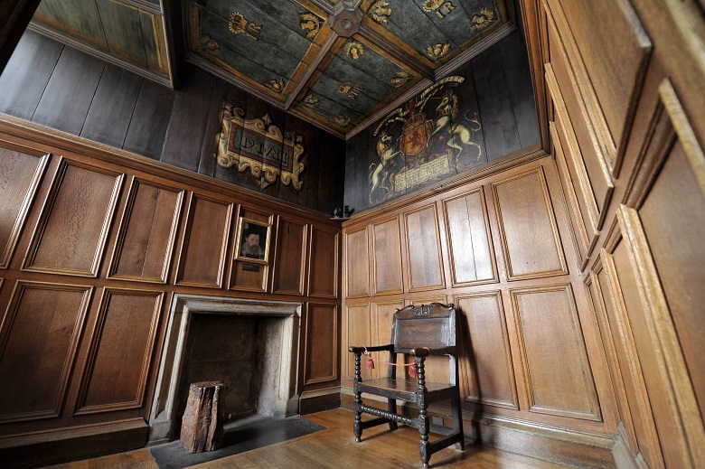 A small room with wooden panelled walls about 2/3 up. Above that the walls are painted black with a coat of arms design on it. There is a small fireplace and a simple chair.