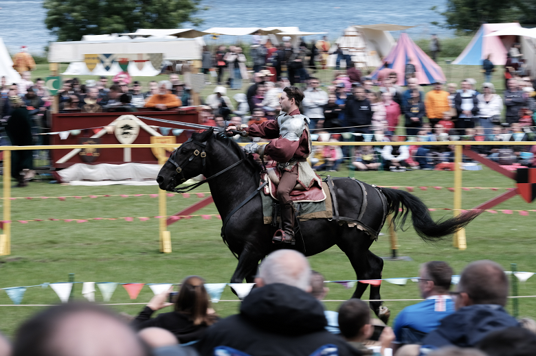 A man dressed in armour charges on a black horse during a jousting match.