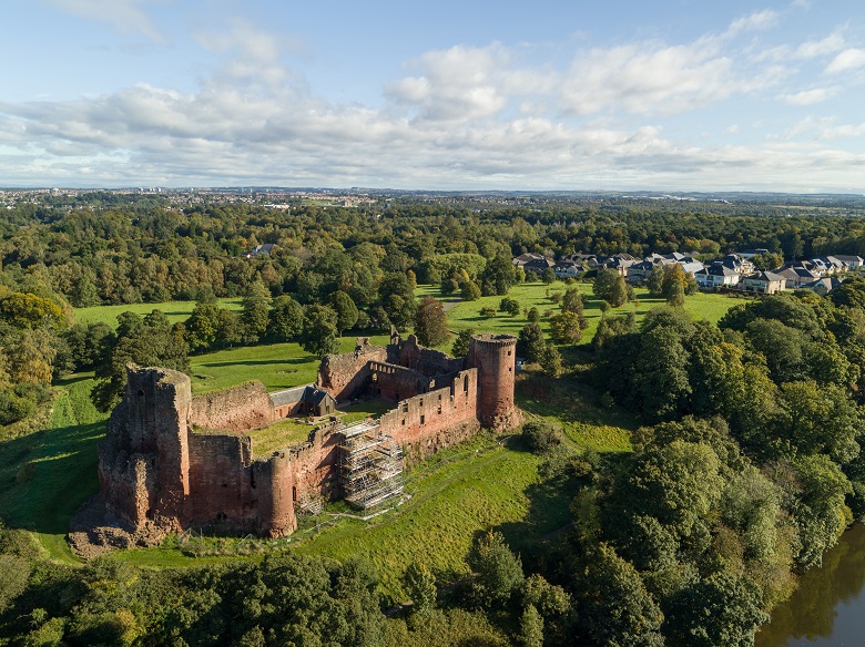 An aerial photo of a large ruined castle close to a river. It has prominent round towers at its corners.