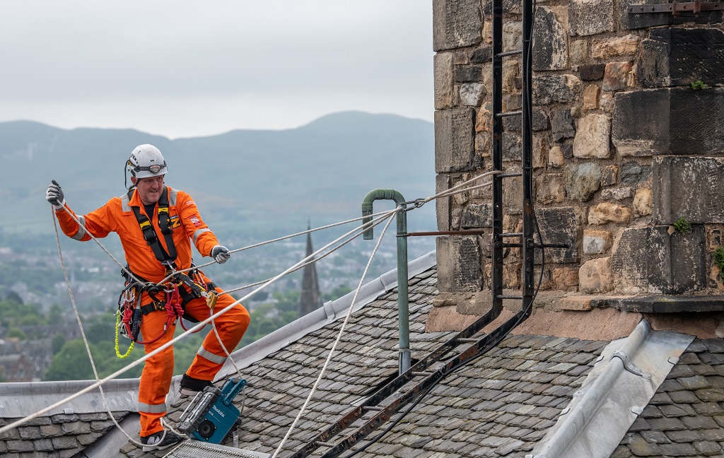 A worker in orange overalls and a hard hat inspects a rooftop at Edinburgh Castle using ropes and a harness.