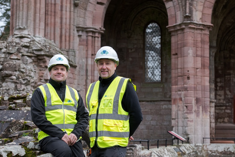 Two workers in yellow hi-vis vests and hard hats pose for a photo in front of the ruins of an abbey
