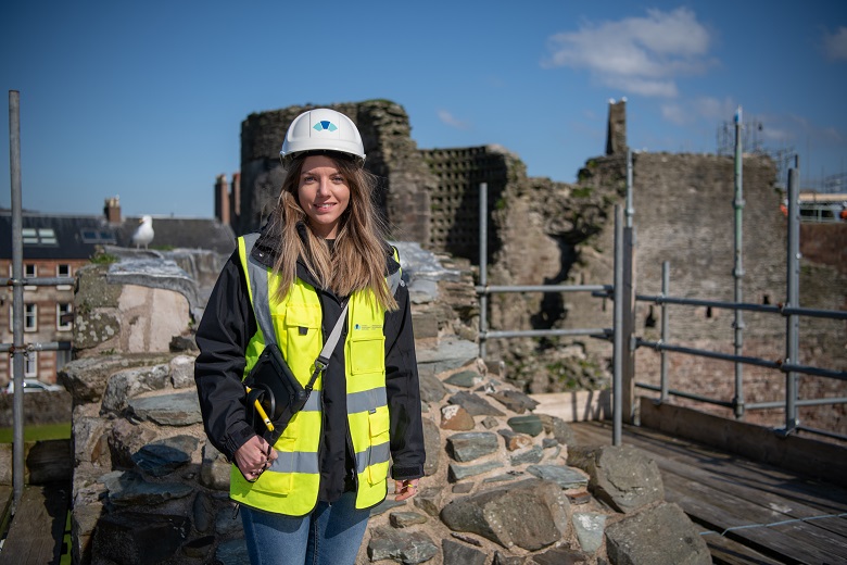 A HES worker wearing a hard hat, a hi-vis vest and holding a clipboard during the inspection of a castle. Part of the ruins are in the background. 