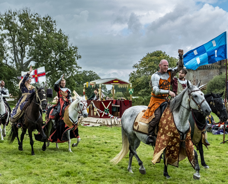 Historical reenactors on horseback at a jousting event. There are jousters in armour and ladies wearing fine dresses. The horses are also dressed up!