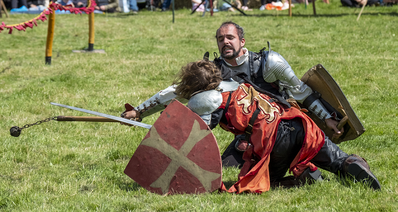 Two costumed reenactors perform a mock fight at a medieval style tournament. At this stage in the fight, both competitors are on the ground, each desperately trying to land a blow on the other. 