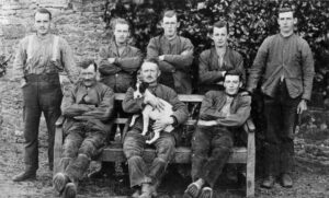 A group of people in a black and white photograph, with three sitting on a bench and five standing in a row behind. They are all wearing farmworkers clothing. The central person seated is holding a black and white terrier dog.