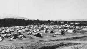 A campsite with dozens of tents and caravans next to a road, with trees and mountains in the distance.