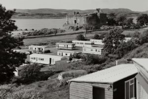 A black and white photo of a castle by a loch, with a series of static caravans in the foreground.