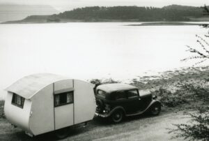 A black and white photo of an old car with a round caravan behind, parked next to a loch.