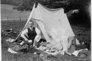 A black and white photo of a person wearing glasses and a waistcoat and tie sitting in front of an open tent. They are surrounded by camping gear, strewn on the grass in all directions.