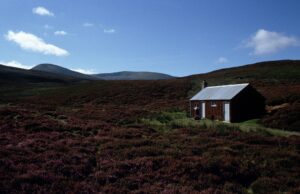 A dark coloured single room hut with two white doors and a chimney in a moorland landscape of heather and mountains.