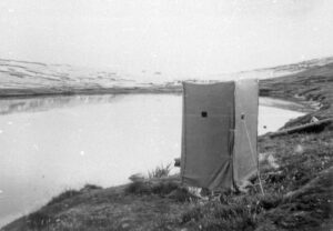 A tent in the shape of an upright rectangle stands next to a loch in a black and white photograph. The tent is made of canvas and has vertical walls, like a box, with a small square hole cut out about halfway up.