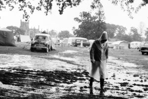 A person wearing a rain mac walks with shoulders slumped out of the rain. In the background is a flooded campsite, tents and cars parked behind an area of very wet grass.