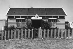 A black and white photo of a single story chalet behind a wooden picket fence. The chalet has two windows, a sloping bitumen roof and a door with a cloud-shaped motif above.